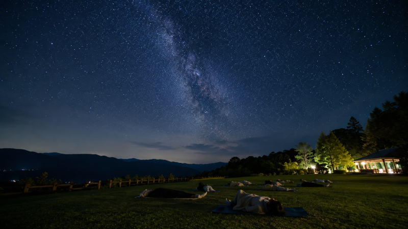 満天の星空の下、マットに横たわり星を眺める人物のシルエット。ヨガ旅(リトリート)の夜に見られる星空の様子。